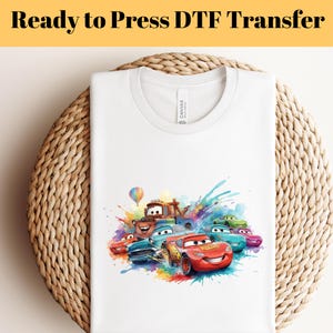 Può includere: T-shirt bianco con una grafica colorata di auto dei cartoni animati, tra cui un'auto da corsa rossa. La maglietta ha il testo "Ready to Press DTF Transfer" in alto. Il design include un effetto arcobaleno.