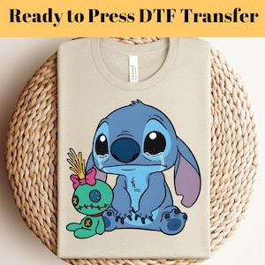 May include: A beige t-shirt with a graphic of the character Stitch from Lilo & Stitch, with a sad expression and tears. The shirt also features a small green doll. The text "Ready to Press DTF Transfer" is at the top.