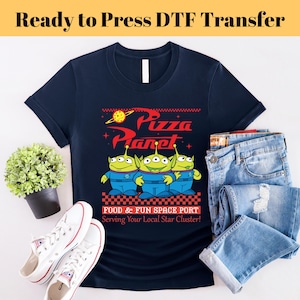May include: Navy blue t-shirt featuring the Pizza Planet logo and three green alien characters. The shirt includes the text "FOOD & FUN SPACE PORT Serving Your Local Star Cluster!" and is paired with blue jeans and white sneakers.