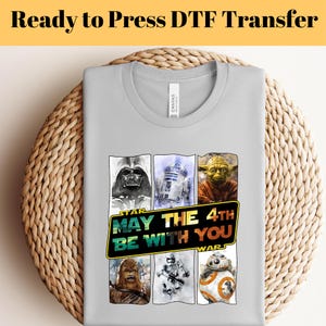 Puede incluir: Camiseta gris claro con un diseño de Star Wars. El diseño incluye a Darth Vader, R2-D2, Yoda, Chewbacca, un Stormtrooper y BB-8. También se muestra el texto "MAY THE 4TH BE WITH YOU".