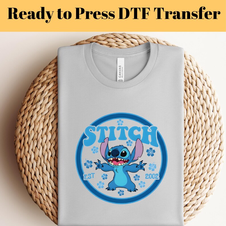 Pode incluir: Uma camiseta cinza claro com um design gr&aacute;fico circular azul e azul claro do personagem Stitch. O design inclui a palavra "Stitch" e o ano "2002". O texto "Ready to Press DTF Transfer" est&aacute; no topo.