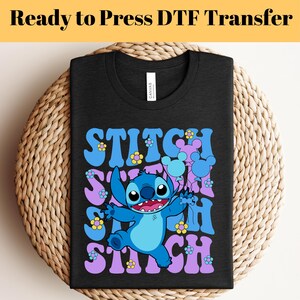 May include: A black t-shirt with a blue Stitch character holding purple balloons with Mickey Mouse ears. The text "Stitch" is repeated in a retro font with purple and yellow flowers.