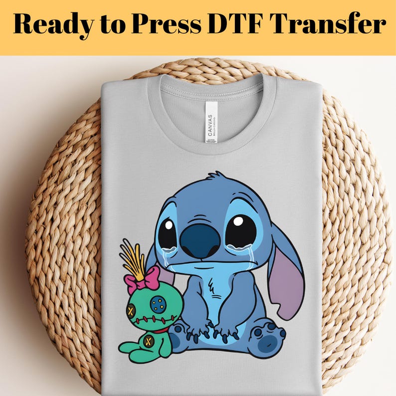 May include: A light gray t-shirt featuring a cartoon image of a blue alien character with large eyes and tears, and a green doll. The text "Ready to Press DTF Transfer" is at the top.