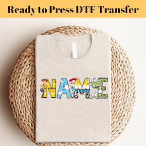 Könnte beinhalten: Ein beiges T-Shirt mit einem DTF-Transfer des Wortes "NAME" in bunten Buchstaben mit Figuren aus dem Toy Story-Film-Franchise.