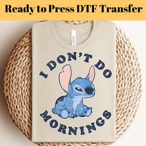 Puede incluir: Camiseta beige con un personaje de dibujos animados azul y el texto "I DON'T DO MORNINGS". El gr&aacute;fico presenta una criatura azul con orejas grandes. La camiseta est&aacute; doblada sobre una superficie tejida. El texto "Ready to Press DTF Transfer" est&aacute; en la parte superior.