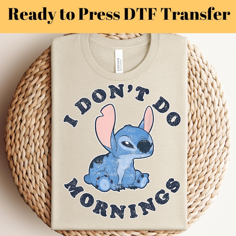 Puede incluir: Camiseta beige con un personaje de dibujos animados azul y el texto "I DON'T DO MORNINGS". El gr&aacute;fico presenta una criatura azul con orejas grandes. La camiseta est&aacute; doblada sobre una superficie tejida. El texto "Ready to Press DTF Transfer" est&aacute; en la parte superior.