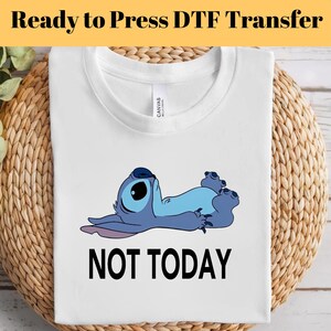 K&ouml;nnte beinhalten: Wei&szlig;es T-Shirt mit einer Cartoonfigur in Blaut&ouml;nen und dem Schriftzug "NOT TODAY" in schwarzer Schrift. Das T-Shirt liegt gefaltet auf einer gewebten Oberfl&auml;che, mit dem Text "Ready to Press DTF Transfer" oben.
