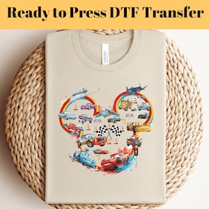 Può includere: T-shirt beige con una grafica colorata di auto e aerei dei cartoni animati disposti a forma di Mickey Mouse. Il testo "Ready to Press DTF Transfer" è in alto. La maglietta è piegata su una superficie tessuta.