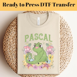 Könnte beinhalten: Beiges T-Shirt mit dem grünen Schriftzug "PASCAL", das ein Cartoon-Chamäleon mit Blumen zeigt. Der Text "Ready to Press DTF Transfer" steht oben. Das T-Shirt liegt gefaltet auf einer gewebten Oberfläche.