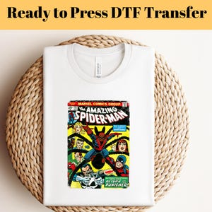 May include: White t-shirt with a vintage comic book print of The Amazing Spider-Man. The comic book cover features Spider-Man in a red and blue suit, fighting a group of villains. The text on the cover reads "The Amazing Spider-Man" and "Return of the Punisher!"
