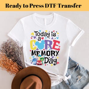以下が含まれることがあります： 「Today is a CORE MEMORY Day」というカラフルなグラフィックがプリントされた白いTシャツ。COREの文字の中にミッキーマウスの頭があり、映画「インサイド・ヘッド」のキャラクターが描かれています。