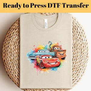 Può includere: T-shirt beige con una grafica colorata di tre auto dei cartoni animati: un'auto da corsa rossa, un'auto classica blu e un carro attrezzi marrone. Il testo "Ready to Press DTF Transfer" è stampato in alto.