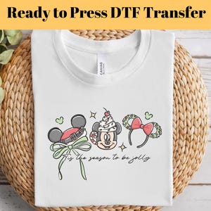 May include: White t-shirt with a festive graphic featuring Mickey Mouse ears and the text "'Tis the season to be jolly." The design includes a Santa hat, a cupcake, and a bow. The shirt is folded on a woven surface.