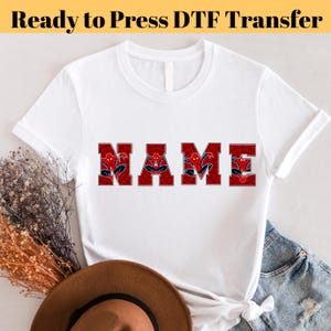 Könnte beinhalten: Weißes T-Shirt mit dem Wort "NAME" in roten Blockbuchstaben, wobei jeder Buchstabe ein Spider-Man-Design zeigt. Der Text "Ready to Press DTF Transfer" befindet sich oben im Bild.