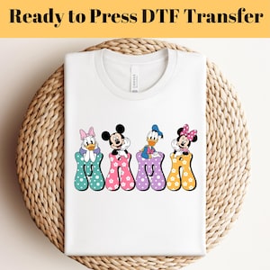 Könnte beinhalten: Weißes T-Shirt mit einem bunten "MAMA"-Design mit Mickey Mouse, Minnie Mouse, Donald Duck und Daisy Duck. Die Buchstaben sind mit Punkten und Blumen verziert.