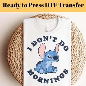 Puede incluir: Camiseta blanca con cuello redondo con un personaje de dibujos animados azul con orejas grandes y el texto "I DON'T DO MORNINGS" en negro. La camiseta se muestra sobre una superficie circular tejida.