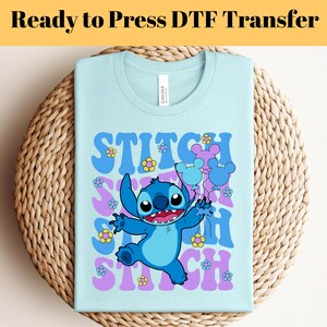 May include: Light blue t-shirt with a blue Stitch character holding balloons. The text "Stitch" is repeated in a retro font with purple and yellow flowers.