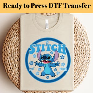 Pode incluir: Camiseta bege com um gr&aacute;fico azul e branco do personagem Stitch. O gr&aacute;fico inclui a palavra "STITCH", o ano "2002" e detalhes florais azuis. O texto "Ready to Press DTF Transfer" est&aacute; na parte superior.