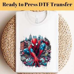 May include: White t-shirt with a vibrant graphic of Spider-Man in a city. The superhero is in his red and blue suit, set against a cityscape. The text "Ready to Press DTF Transfer" is at the top of the image.