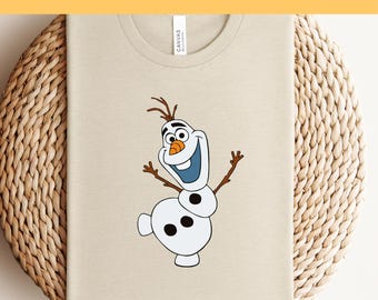 Puede incluir: Camiseta beige con una imagen de dibujos animados de Olaf, el muñeco de nieve de Frozen. El texto "Ready to Press DTF Transfer" está en la parte superior. La camiseta está sobre una superficie circular tejida.