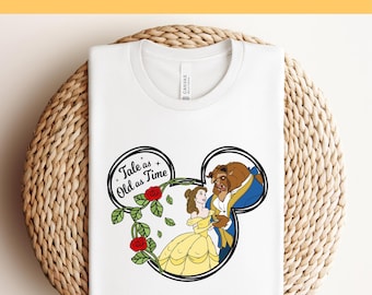 Könnte beinhalten: Weißes T-Shirt mit einem schwarz-weißen Design von Belle und dem Biest aus Die Schöne und das Biest. Das Design hat die Form eines Mickey-Mouse-Ohrs und enthält den Text "Tale as Old as Time".