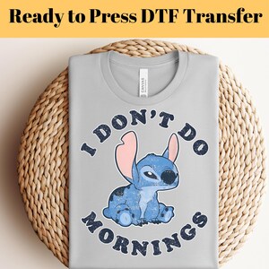 Puede incluir: Camiseta gris claro con una imagen de dibujos animados de un extraterrestre azul con orejas grandes y el texto "I DON'T DO MORNINGS". La camiseta est&aacute; doblada sobre una cesta tejida.