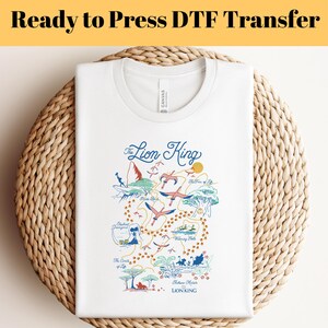 May include: White t-shirt with a colorful graphic of a map of the Lion King movie. The map includes locations like Pride Rock, the Watering Hole, and the Elephant Graveyard. The text "The Lion King" is written in a cursive font at the top of the graphic.