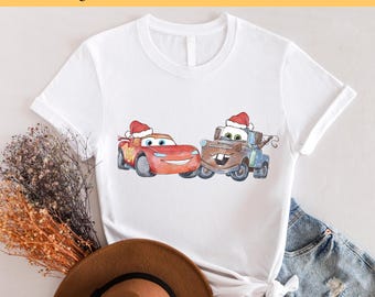 May include: White t-shirt with a red and rusty colored cartoon image of two cars wearing Santa hats. The cars are from the Disney Pixar movie Cars. The text "Ready to Press DTF Transfer" is at the top of the image.
