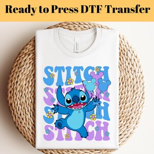 May include: White t-shirt with a blue Stitch character holding balloons with Mickey Mouse ears. The text "Stitch" is repeated in a purple and blue retro font with yellow flowers.