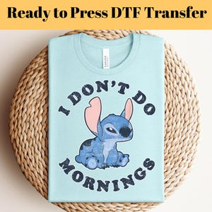 Puede incluir: Una camiseta azul claro con el texto "I DON'T DO MORNINGS" y un personaje de dibujos animados. La camiseta est&aacute; doblada sobre una superficie tejida. Las palabras "Ready to Press DTF Transfer" est&aacute;n en la parte superior.