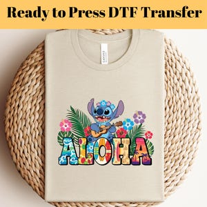 Puede incluir: Una camiseta beige con un gráfico colorido de un personaje de dibujos animados azul tocando el ukelele, rodeado de flores tropicales y la palabra "ALOHA". La camiseta está doblada sobre una superficie tejida.