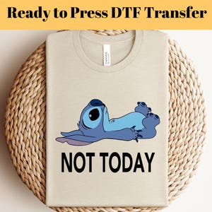 K&ouml;nnte beinhalten: Beiges T-Shirt mit einer Cartoonfigur in Blau und dem schwarzen Text &bdquo;NOT TODAY&ldquo;. Das T-Shirt liegt gefaltet auf einer gewebten Oberfl&auml;che. Der Text &bdquo;Ready to Press DTF Transfer&ldquo; steht oben.