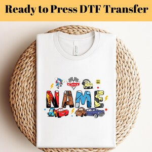 Könnte beinhalten: Weißes T-Shirt mit einem farbenfrohen Cars-Design, das das Wort "NAME" integriert. Das Design beinhaltet Lightning McQueen, Mater und andere Figuren aus dem Film.