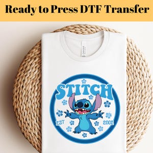 Pode incluir: Camiseta branca com um gr&aacute;fico azul do Stitch. O design inclui a palavra "Stitch" em um c&iacute;rculo azul, o personagem Stitch e o texto "EST 2002". A camiseta &eacute; exibida em uma superf&iacute;cie tecida. O texto "Ready to Press DTF Transfer" est&aacute; no topo.