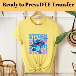 May include: Yellow short-sleeved t-shirt with a blue Stitch character graphic. The graphic features Stitch holding two purple balloons with Mickey Mouse ears. The text "STITCH" is repeated in a retro font in purple and pink.