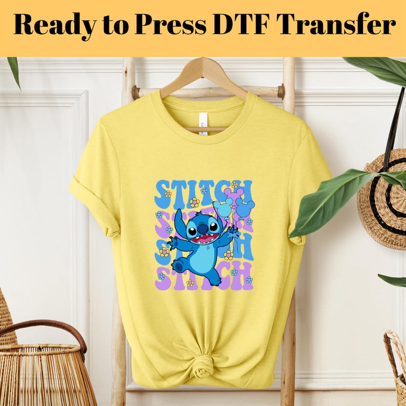 May include: Yellow short-sleeved t-shirt with a blue Stitch character graphic. The graphic features Stitch holding two purple balloons with Mickey Mouse ears. The text "STITCH" is repeated in a retro font in purple and pink.