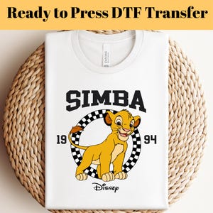 以下が含まれることがあります： ディズニーのライオンキングのシンバのグラフィックが特徴の白いTシャツ。デザインには、太字の黒い文字で「SIMBA」という単語、数字の「1994」、シンバの漫画の画像が含まれています。シャツは織られた表面に表示されます。上部には「Ready to Press DTF Transfer」というテキストがあります。