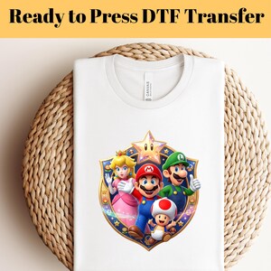 May include: White t-shirt featuring a colorful graphic of Mario, Luigi, Princess Peach, and Toad. The design is set within a gold and blue shield shape. The text "Ready to Press DTF Transfer" is at the top.