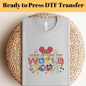 Könnte beinhalten: Ein hellgraues T-Shirt mit dem Text "Ready to Press DTF Transfer" oben. Das Shirt zeigt eine farbenfrohe Grafik von Mickey Mouse und seinen Freunden mit den Worten "Drink Around The World Tour".