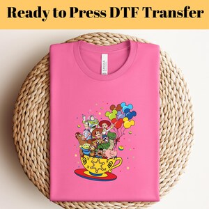 Pode incluir: Camiseta rosa com um gr&aacute;fico dos personagens do Toy Story em uma x&iacute;cara de ch&aacute; com bal&otilde;es. Os personagens incluem Woody, Buzz Lightyear, Jessie, Rex e Hamm. O texto "Ready to Press DTF Transfer" est&aacute; na parte superior da imagem.