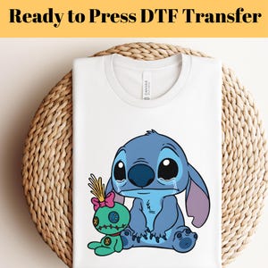 May include: White t-shirt with a blue cartoon character and a green doll graphic. The text "Ready to Press DTF Transfer" is at the top. The shirt is displayed on a woven, circular surface.