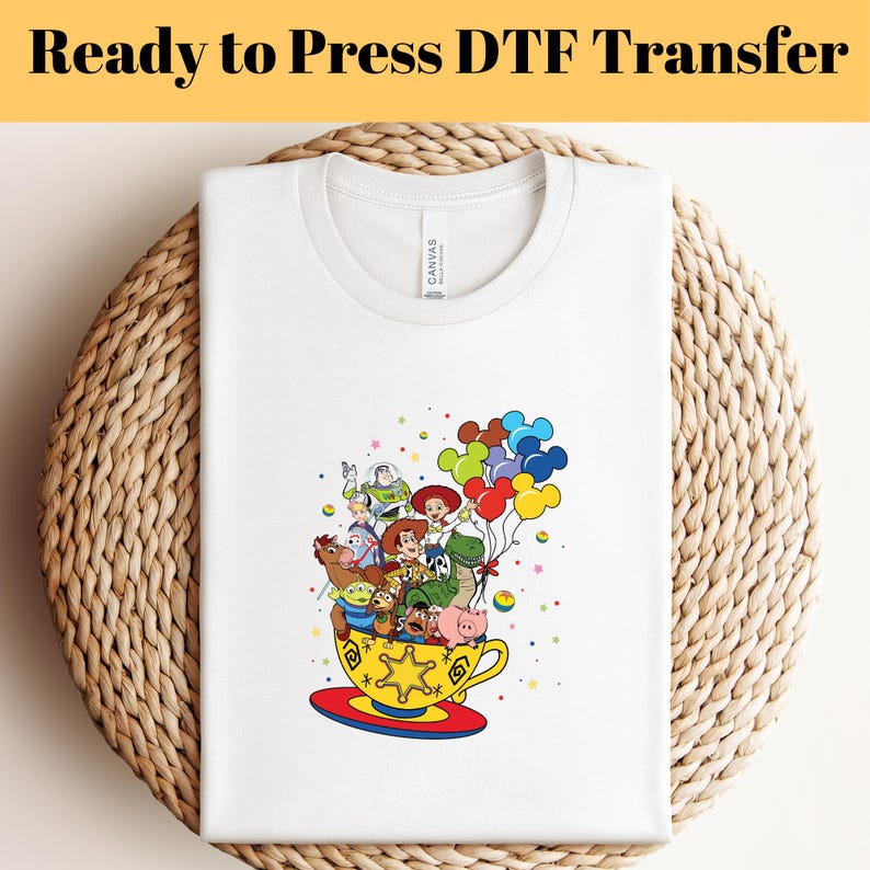 Pode incluir: Camiseta branca com um gr&aacute;fico colorido dos personagens do Toy Story em uma x&iacute;cara de ch&aacute; com bal&otilde;es. O texto "Ready to Press DTF Transfer" est&aacute; na parte superior da imagem.