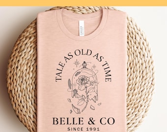 Könnte beinhalten: Hellrosa T-Shirt mit Rundhalsausschnitt. Das Shirt zeigt eine schwarze Grafik einer Rose in einer zerbrochenen Glaskuppel, mit dem Text "TALE AS OLD AS TIME" und "BELLE & CO SINCE 1991".