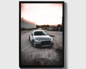 May include: A silver Audi TT-RS parked in a paved lot with a brick wall in the background. The car is facing the camera and the sky is a light pink color.