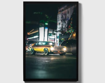May include: A yellow and black classic car driving down a city street at night. The car has a black interior and a black hood. The car is driving past a building with lights on.