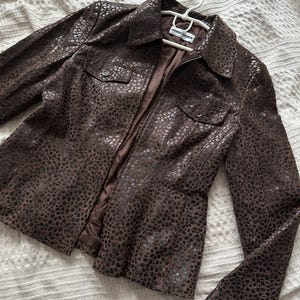 May include: A brown leather jacket with a spotted pattern, featuring a collared neckline, two chest pockets, and a zipper closure. The jacket is displayed on a white hanger.