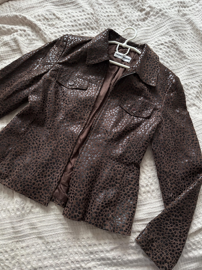 May include: A brown leather jacket with a spotted pattern, featuring a collared neckline, two chest pockets, and a zipper closure. The jacket is displayed on a white hanger.