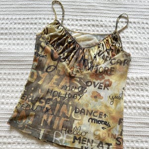 May include: A sleeveless camisole top featuring a gathered V-neckline and thin spaghetti straps. The fabric has a yellow, brown, and tan tie-dye effect with various black printed words and phrases including "never," "holiday," "dance," and "men at."