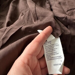 May include: A brown garment with a tag held by a hand. The tag has text in multiple languages, including "GERRY WEBER" and fabric composition details. The garment's fabric appears to be a dark brown color.