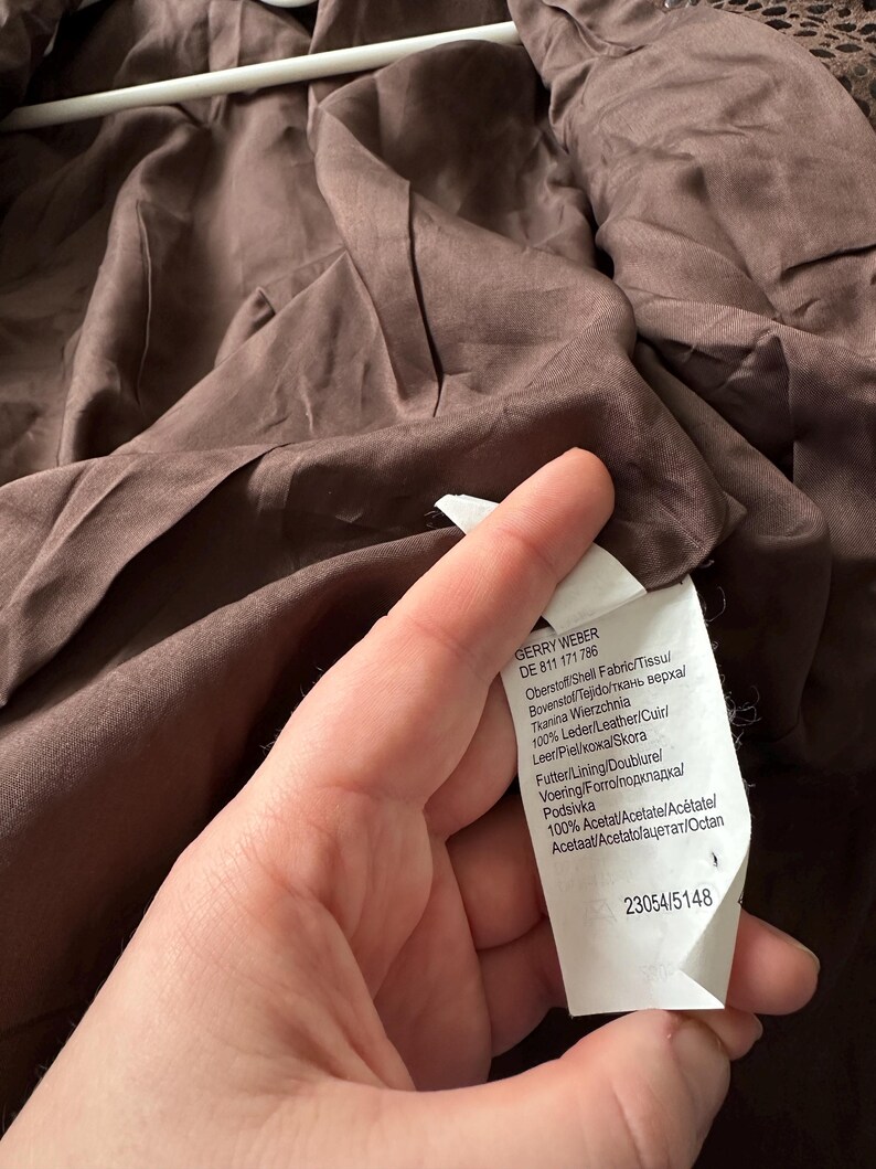 May include: A brown garment with a tag held by a hand. The tag has text in multiple languages, including "GERRY WEBER" and fabric composition details. The garment's fabric appears to be a dark brown color.